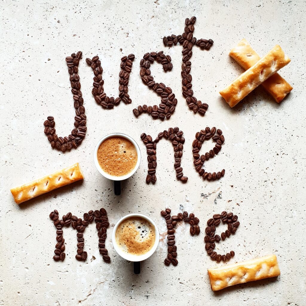 Two cups of coffee sit among coffee beans arranged to spell "just one more," with three pieces of pastry placed around them, creating a cozy summer special scene on a light surface. Top Dose Roasters
