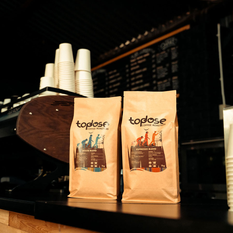 Two brown bags of Topdose Coffee Roasts, House Blend and Espresso Blend, from top coffee roasters, are displayed on a counter beside a stack of paper cups and a coffee machine. Top Dose Roasters