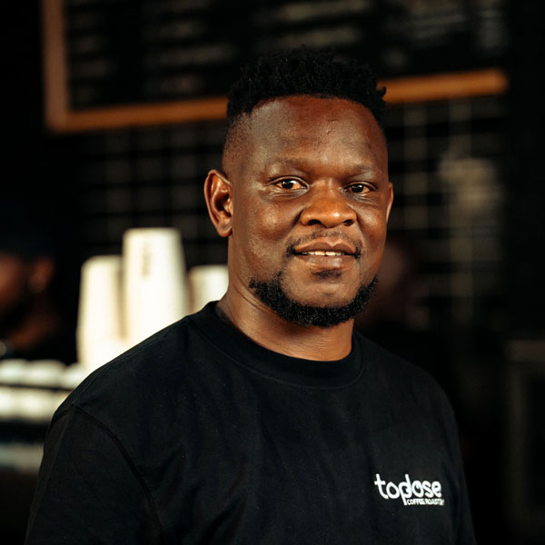 Neo Mkhethoni – Founder & Head Barista wearing a black "topdose" t-shirt stands indoors, with coffee cups and a menu board visible in the blurred background. Top Dose Roasters